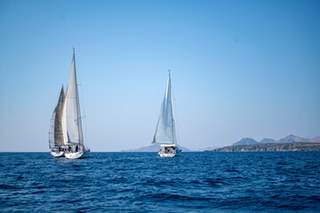 Fototapeta premium Luxury sailing yachts and white boats cruising on the deep blue Mediterranean sea during an active summer regatta ocean vacation with a scenic coastal resort town and mountains in the background