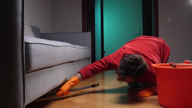 Person wearing an orange shirt and gloves scrubbing hard to reach areas of a wooden floor under a couch with a mop near a cleaning bucket