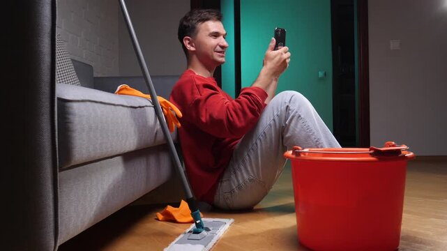 Young man sitting on the floor, laughing and watching a video on his smartphone, avoiding cleaning activities with a mop and bucket nearby in a living room