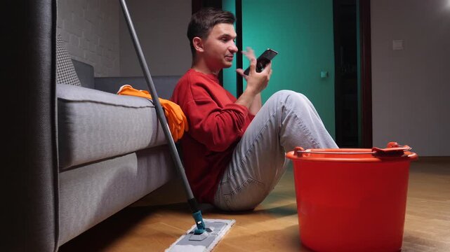 Frustrated man pausing during house cleaning, sitting on the floor with a mop and bucket, gesturing with phone, experiencing stress from household chores