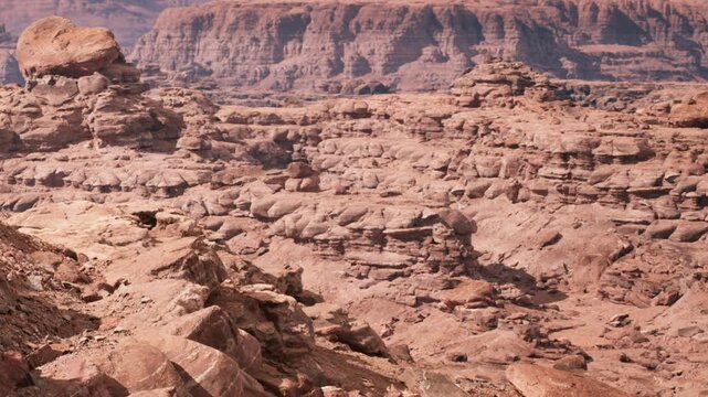 Large rock formations in a dry desert area. The ground is made of different layers of stone and sand. The sky is clear above the rugged terrain.