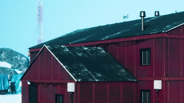 A red building stands in a snowy area surrounded by mountains. Some equipment is visible near the building. The sky is clear, and there is little vegetation in the area.
