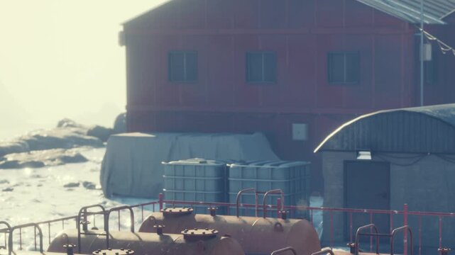 A red building stands next to the water with storage tanks in front. The sun sets in the background, creating a warm glow on the rocks. The scene shows a quiet moment at the shore.