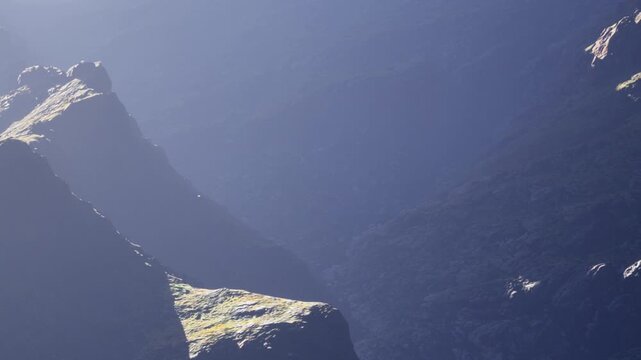 A wide landscape shows tall mountains and deep valleys. Sunlight shines on rocky surfaces. The scene appears untouched and quiet with little vegetation scattered around.