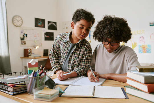 Medium shot of Black teenager helping younger boy studying and writing in notebooks at desk, supporting home learning and sibling education. Useful for tutoring, parenting, academic support marketing