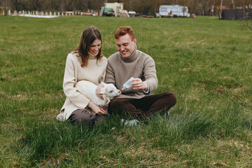 Fototapeta premium Couple sitting on green grass in a park, a white kid goat between them, sharing a gentle moment of care and play, cozy sweaters, casual lifestyle, and rustic charm.