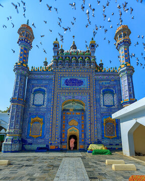 View of the Shrine of Sachal Sarmast with its intricate blue tile work and minarets under a clear sky, with pigeons flying overhead in Daraza Sharif, Sindh, Pakistan.
