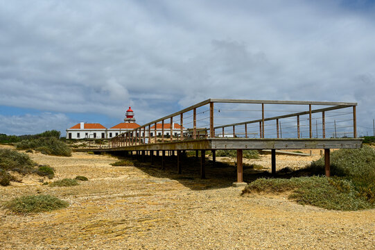 View of a lighthouse with a red top and white buildings, situated behind a wooden observation deck on a dry, grassy landscape under a cloudy sky in Algarvia, Azores, Portugal.
