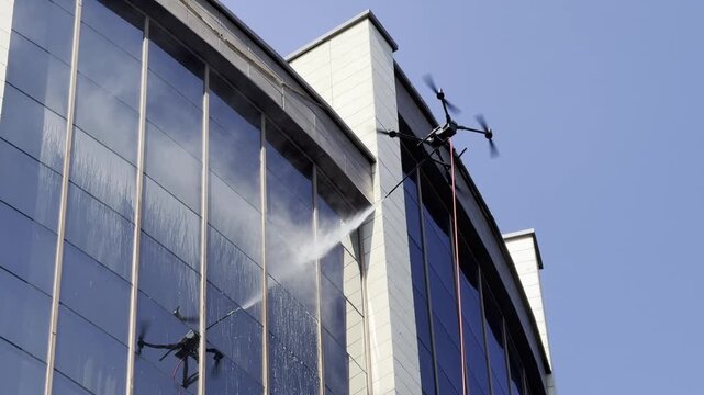 Drone is washing windows. Drone spraying water from a suspended nozzle onto upper windows of a modern building. Red hose hangs below the aircraft against blue sky, showcasing facade cleaning