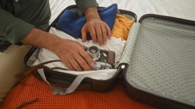 Man packing a hard shell suitcase on a bed, hands adjusting straps and placing a vintage camera and sunglasses in the case in building; anticipation travel preparation.
