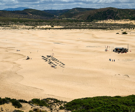 View of a vast sandy beach with rows of straw umbrellas, dunes, and a small beach structure under a cloudy sky in Algarvia, Azores, Portugal.