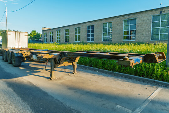 Long metal container chassis trailer with multiple axles parked on a street beside a low industrial brick building and grassy verge. The rusty steel frame