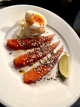 Close-up of smoked salmon slices served with a soft-boiled egg, lime wedge, and sesame seed garnish on a white plate. Fresh seafood dish ideal for brunch, breakfast, healthy eating, gourmet dining, pe