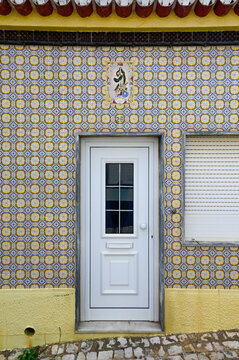 View of a traditional house facade decorated with intricate blue and yellow azulejos tiles, featuring a religious icon plaque above a white door in Algarvia, Azores, Portugal.