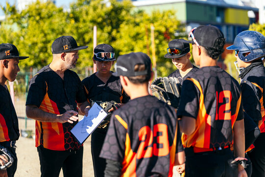 Coach holding a clipboard and talking with young baseball players in a huddle, discussing game strategy on a field
