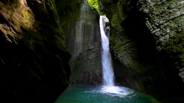 Hidden Kozjak waterfall drops into a turquoise pool inside a dark mossy gorge, framed by steep rock walls and natural cave shadows.