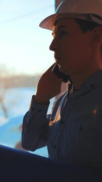 A man wearing a hard hat is talking on a phone. The man stands near a large window, with sunlight illuminating the snowy landscape outside.