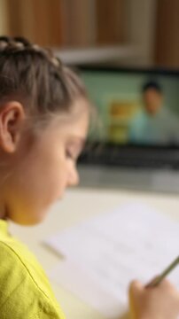 A girl focuses on her writing while sitting at a table. A screen in the background shows a colorful animation that the girl observes.
