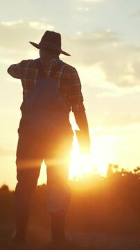 A man stands in a field during sunset, silhouetted against the sky. The man wears a hat and overalls, reflecting a typical farming lifestyle amidst the glowing sun.