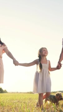 A woman and a man hold hands with a girl in a field. The girl enjoys the moment while the sun sets in the background, creating a calm atmosphere.