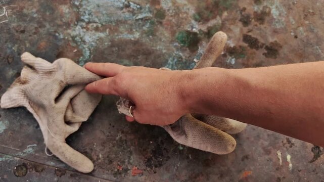 A woman's hand picks up a pair of worn gardening gloves from an old rusty copper table outdoors, gardening equipment, manual labor, protective gear, outdoor work, rustic lifestyle