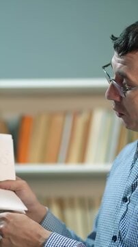 A man examines a book intently. The man holds the book while sitting in front of a shelf filled with various books.