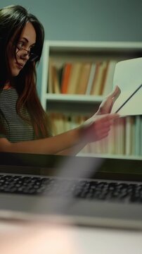 A woman with long hair and glasses is presenting a document. The woman gestures towards the paper while seated at a desk with a laptop in an organized room.