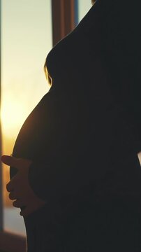 A pregnant woman stands by a window, silhouetted against a sunset. The woman gently cradles her belly, highlighting the beauty of motherhood during this intimate moment.