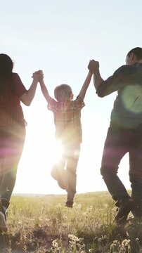 A woman and a man hold hands with a boy in a field. The boy joyfully swings between the woman and man against a bright sunlight backdrop.