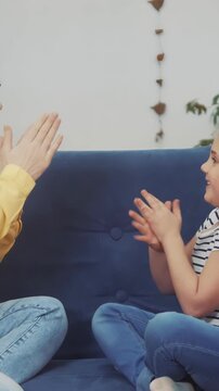 A woman and a girl engage in a playful game on a blue couch. Both the woman and the girl smile and clap their hands in rhythm during the playful interaction.