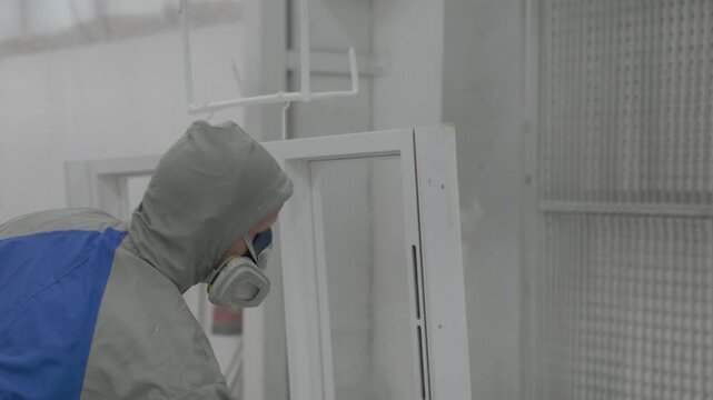 Worker in protective suit spray painting a wood frame window inside an industrial factory booth with paint containers and ventilation wall