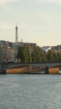 Vertical view of a classic stone bridge arching over the Seine River in Paris, with historic Parisian buildings and the iconic Eiffel Tower rising elegantly into the soft afternoon sky.