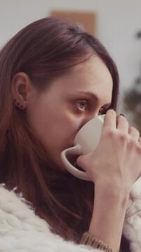 A woman with long hair is sipping from a cup in a cozy setting. The woman appears thoughtful while holding the cup in her hands, enjoying her drink.