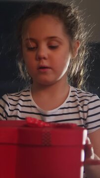 A girl with long hair examines a red gift box. The girl is focused on the red box as she opens it, revealing her curiosity about the contents inside.