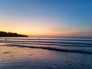 Fototapeta premium Tropical beach at twilight with orange and blue sunset sky, silhouetted headland and fishing boat on calm ocean