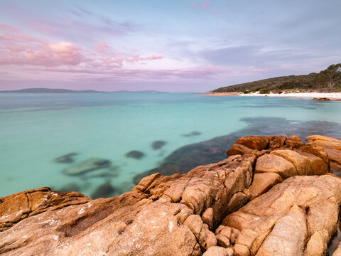 View of Whalers Cove with turquoise water, rocky foreground, and a sandy beach under a soft sunset sky in Albany, WA, Australia.