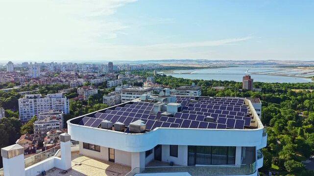 Solar panels are installed on a building roof in the city. Below, the urban area and river are visible in the background. The scene shows clear weather with daylight.