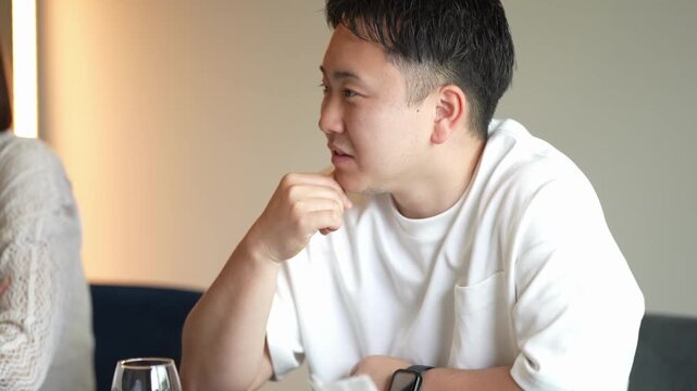Man in white shirt listens intently during conversation. He rests hand near mouth, thoughtful expression. Setting suggests relaxed, intimate dining encounter. Ideal for lifestyle, relationship