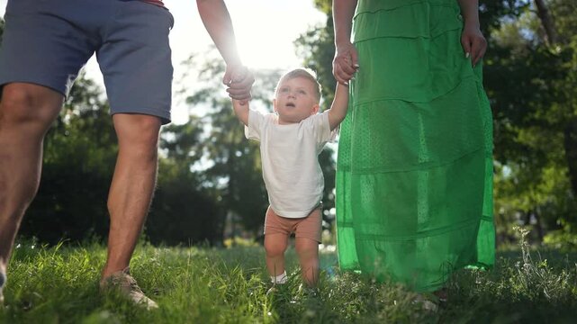 Child learn to walk with mom and father. Parents hold child hands while guiding first steps in park. Family supports child as learn to walk. Mom and father help child develop walking skills outdoors.