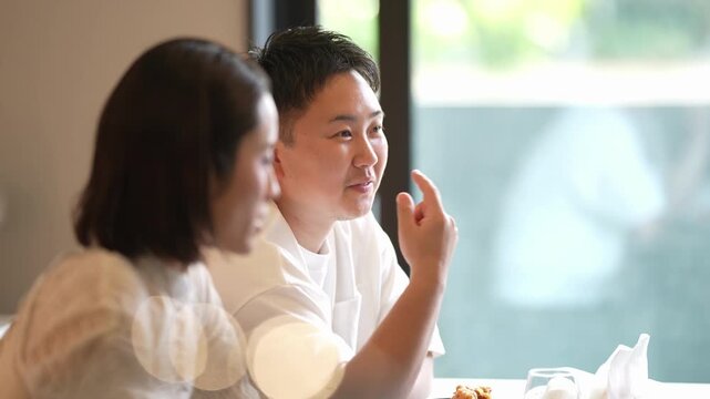 Two people share a quiet, relaxed meal. Soft light filters through large windows behind them. The woman rests her head, smiling gently. The man leans forward, amused by something unseen