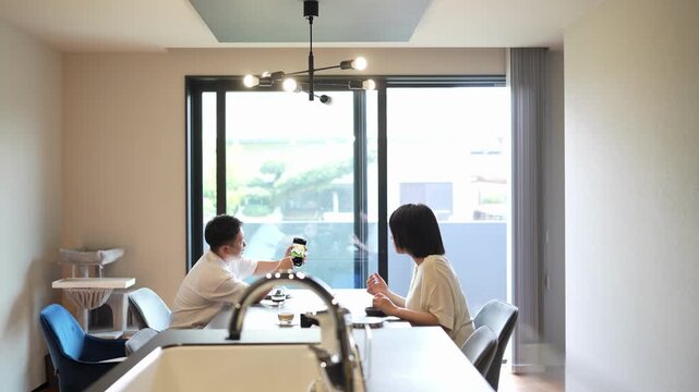 Soft natural light fills minimalist kitchen space. Couple shares quiet moment over drinks at sleek island counter. They smile gently, enjoying each other's company. Ideal for cozy home, romantic date