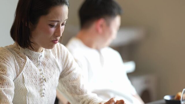 Woman gestures mid-conversation at a cafe. She wears a lace blouse and smartwatch. Her expression shows thoughtful engagement. Background man appears focused, blurred. Ideal for business, casual