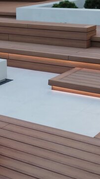Vertical video. Wooden terrace steps with LED lighting beside a modern patio and barbecue area.