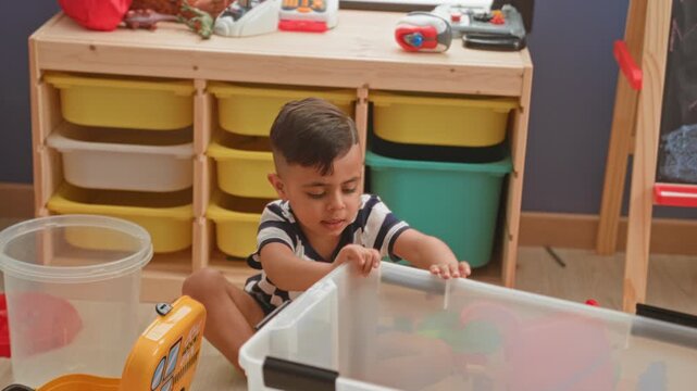 Little boy sits on the floor playing with a yellow toy truck and clear plastic bin in a playroom building; quiet concentration.