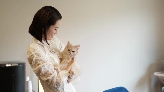 Young woman smiles warmly while holding a kitten. Soft light highlights her gentle expression. Her arms cradle the fluffy, cream-colored feline. The kitten looks up, curious and relaxed