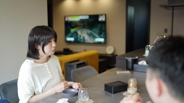Two people sit at a modern table. They watch a racing game on a TV screen. The woman looks engaged, thoughtful. The man's attention is fixed on the game. Their setting feels relaxed and contemporary