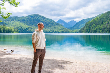 Fototapeta premium Bald young man wearing glasses, white hoodie, plaid trousers, Travel and tourism stylish clothing choices, pebble beach, turquoise lake, mountains, forest, summer vacation, travel, fashion portrait