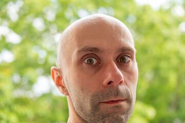 Naklejka premium Close up outdoor portrait bald man, middle aged male, shaved head, green blurred nature background, outdoor lifestyle, calm expression inner peace, serious face, natural light sunny day