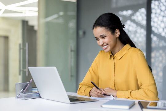Smiling young businesswoman working diligently at her modern office desk, actively engaging with her laptop and writing notes, embodying productivity and success in her professional career