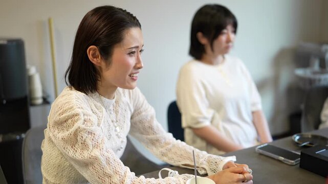 Two women sit at a desk in an office. One smiles warmly, hands clasped together. The other looks forward, expression calm and attentive. They appear to be in a meeting or collaborative discussion